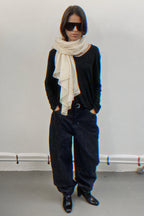 A model stands against a plain light gray wall wearing a black long-sleeve top, loose dark jeans, black pointed shoes, a cream cashmere scarf, and black sunglasses.