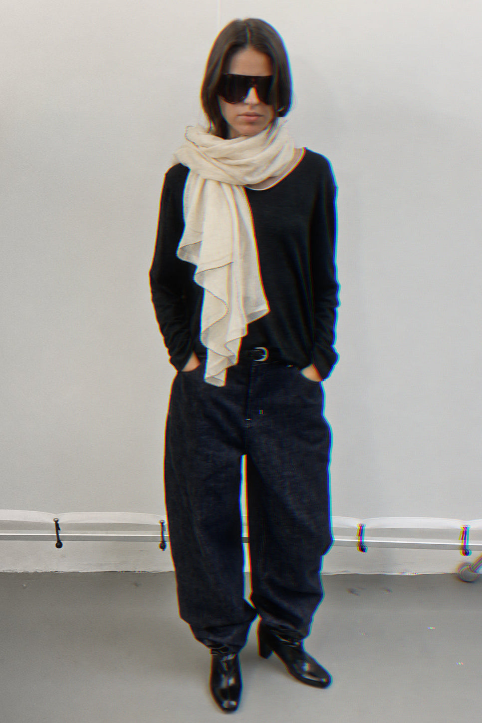 A model stands against a plain light gray wall wearing a black long-sleeve top, loose dark jeans, black pointed shoes, a cream cashmere scarf, and black sunglasses.