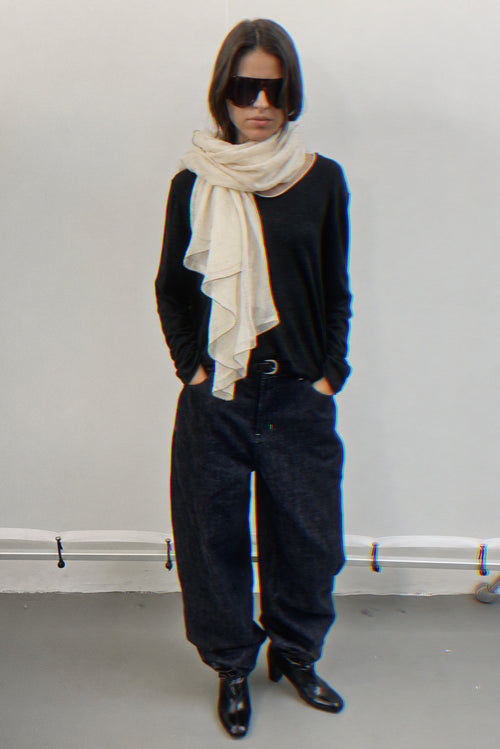 A model stands against a plain light gray wall wearing a black long-sleeve top, loose dark jeans, black pointed shoes, a cream cashmere scarf, and black sunglasses.