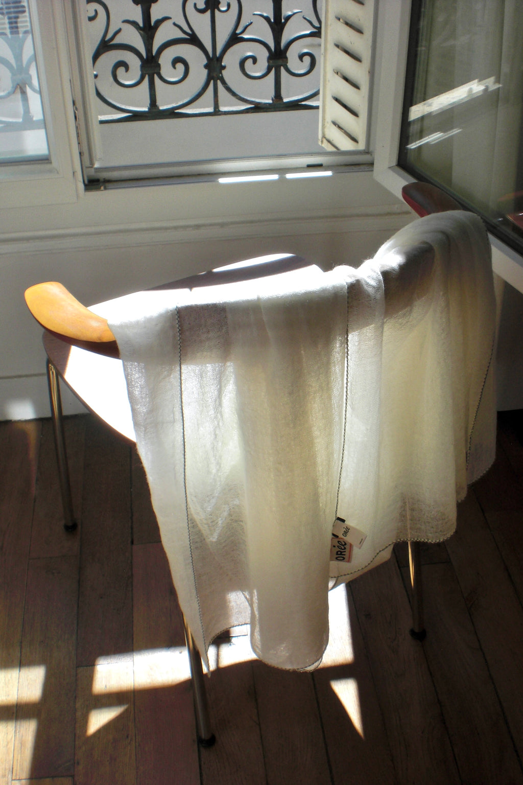 A sheer cream-colored cashmere scarf draped over a wooden chair in a sunlit parisien room with hardwood floors and a window.